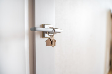 Keys with a keychain in the form of a house hang on the handle of an open white door
