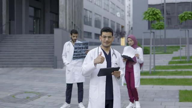Fornt view of smiling young arabic male doctor with tablet pc, standing outdoors near hospital building, showing thumb up. Two diverse colleagues standing behind him and working with xray
