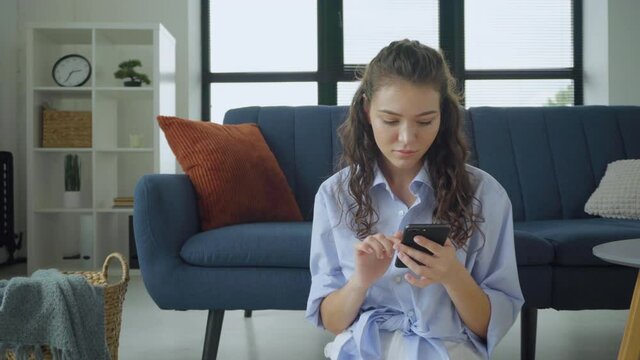 Young woman texting on smartphone while sitting on the floor in the living room. Smiling curly girl reading a message, browsing on internet and using app, having fun conversations chat with her mobile