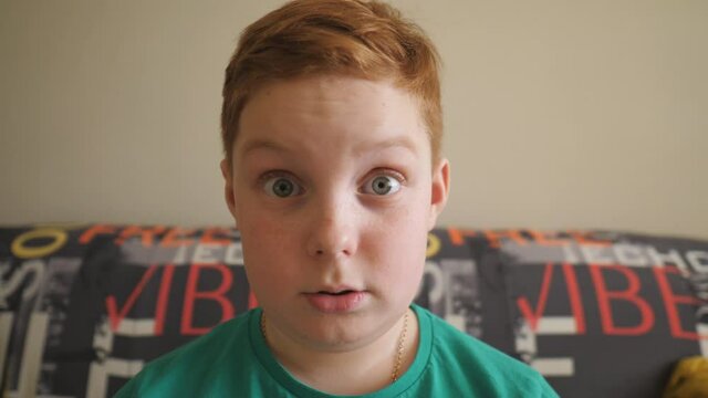 Little red-haired boy sitting on the sofa and looking into camera with shocked expression. Close up scared male face of small child. Portrait of ginger kid with freckles showing fright indoor. Slow mo