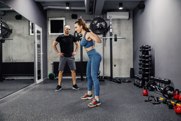 Fototapeta na wymiar Barbell training with weights. A young woman, in sportswear and in good shape, does squats and holds a barbell on her back. In the background is an individual fitness trainer who is support her