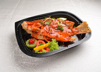 deep fried crispy whole grouper fish seafood with sweet and sour chilli sauce and lychee fruits on top in black bento tray box for asian take away halal menu