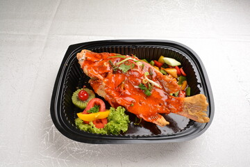 deep fried crispy whole grouper fish seafood with sweet and sour chilli sauce and lychee fruits on top in black bento tray box for asian take away halal menu