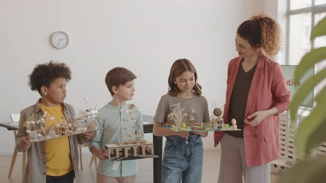 Medium Long POV Of Young Nice Caucasian Teacher Asking Her Multiethnic Elementary School Students About Their Projects, All Standing In Classroom Then Smiling And Waving Hands On Camera