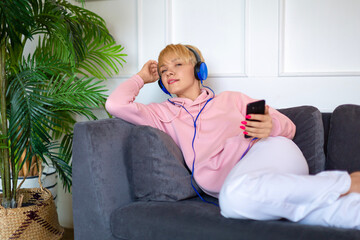 A young woman sits on the couch with a tablet and listens to music through headphones.