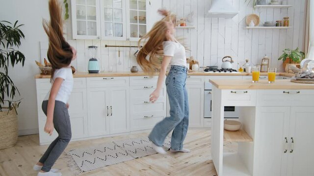 Happy girl teenager child schoolgirl daughter having fun with beloved single mom adult woman mother babysitter with long hair dancing in home kitchen jumping to music spinning like airplane laughing