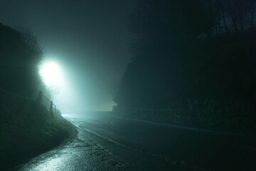 A spooky, eerie country road passing through a hill. On a mysterious foggy, winters night. With a street light in the distance.