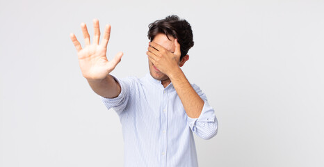 hispanic handsome man covering face with hand and putting other hand up front to stop camera,...