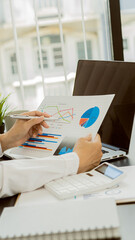 A young financial market analyst works in the office on his laptop while sitting at a wooden table. Businessman analyzing documents in hand Graph and financial concept calculator with vertical images.