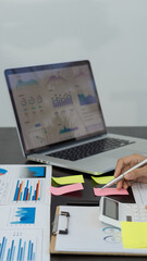 A young financial market analyst works in the office on his laptop while sitting at a wooden table. Businessman analyzing documents in hand Graph and financial concept calculator with vertical images.