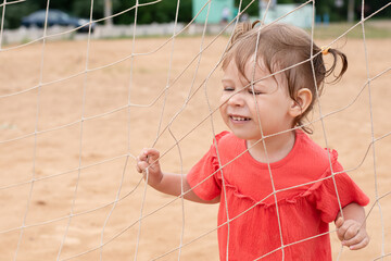 Emotional child plays in the sand by the sea. The girl plays at the football goal on the beach. The baby is holding onto the net of the soccer goal.