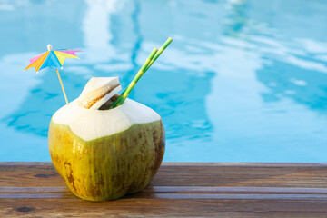 Fresh  green coconut drink with paper straw  and rainbow umbrella  standing on wooden board  near pool  water  tropical beach resort background  with copy space Vacation  in exotic  resort  concept .