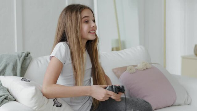 Cheerful Caucasian schoolgirl teenager happy active girl child daughter playing video game online alone at home sitting on couch play console holding joystick controller emotionally gaming videogame