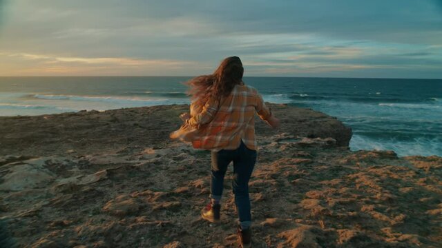 Cinematic wanderlust lifestyle young woman excited run to edge of cliff, jump and shout into wind and sunset. Inspiring travel adventure, forever young and freedom concept. Female traveller at sunset