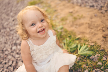 Fototapeta na wymiar Little funny cute blonde girl child toddler with curls in white dress and with mud on her face sits on the ground on the street outside at summer. Baby laughing. Happy healthy childhood concept