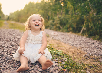 Little funny cute blonde girl child toddler with curls in white dress and with mud on her face sits on the ground on the street outside at summer. Baby laughing. Happy healthy childhood concept