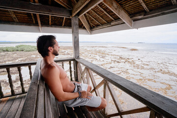 Relaxed and cheerful. Happy young bearded man resting on terrace near the sea.