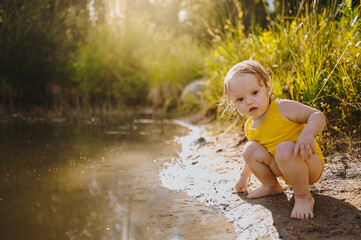 Little funny cute blonde girl child toddler in yellow wet bodysuit playing by the lake waterside shore at sunset outside. Baby with mud on hands and legs. Water activities at summer