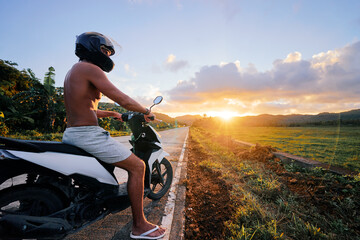 Tropical travel and transport. Man in helmet riding scooter on the empty road with sunset landscape.