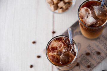 Iced coffee in a tall glass with cream or milk and beans, straws on light background. Cold tasty summer refreshment beverage concept. Selective focus, copyspace. Flat lay, top view.