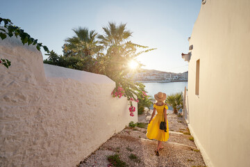 Young traveling woman in hat and yellow dress walking on old town enjoying the sea view. - obrazy, fototapety, plakaty
