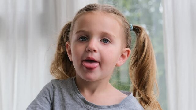 Funny preschooler girl blonde with long pigtails and blue eyes shows tongue pulling face and looks straight at home against window closeup