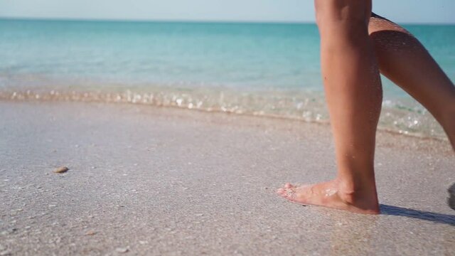 Slim Girl Legs Walking along the Sandy Beach along the Transparent Turquoise Sea. Unrecognizable Pretty Woman Walks at Seaside Surf. Summer Vacation and Travel. Slow motion.