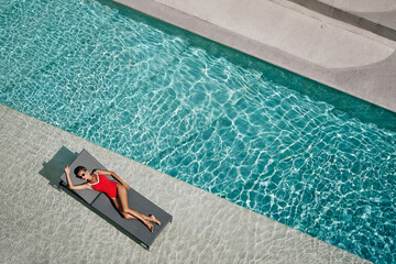 Enjoying suntan. Vacation concept. Top view of slim young woman in red swimwear on the sun lounger near the swimming pool.