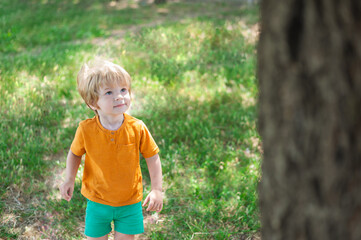 little blond boy smiles, walks on green grass in summer, looks at a tall tree