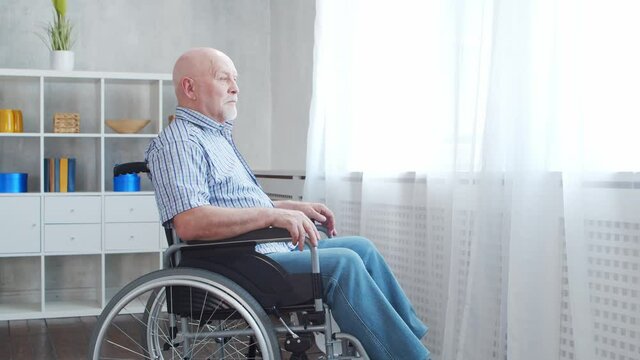 Disabled old man is sitting in a wheelchair at home alone. A handicapped person in a wheelchair is looking out the window. Paralysis and disability.