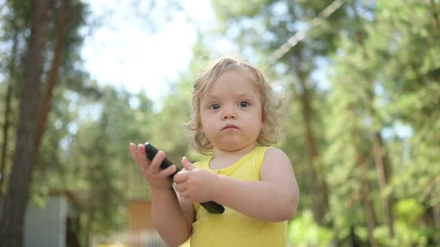 Little funny cute blonde girl child toddler holding playing big black smartphone outside at summer at countryside. Healthy happy childhood concept. Kids and technologies