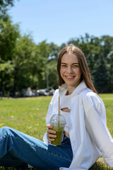 A young girl is sitting in the park on the grass and drinking a green smoothie