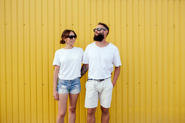 Young smiling pretty happy cheerful positive couple in blank white t-shirts near color background on street. Mockup for design.