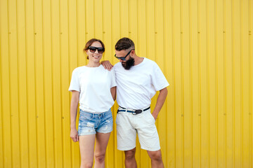 Young smiling pretty happy cheerful positive couple in blank white t-shirts near color background on street. Mockup for design.