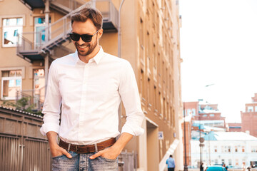 Portrait of handsome confident stylish hipster lambersexual model.Modern man dressed in white shirt. Fashion male posing in the street background near skyscrapers in sunglasses
