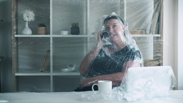 Self isolation. Social anxiety. Personal barrier. Low immunity. Worried female office worker covered with polyethylene film talking on phone in light workspace.