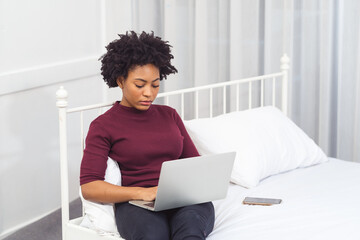 Happy casual beautiful american african woman working on laptop computer while setting  on the bed in the house.