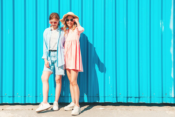 Two young beautiful smiling hipster female in trendy summer clothes and dress.Sexy carefree women posing in the street near blue wall.Positive pure models having fun at sunset, hugging and going crazy