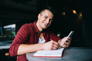 Portrait of cheerful male student smiling at camera while sitting at cafeteria table with education textbook for studying and smartphone technology for informative online research, mobile using