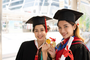 Two Asian women who wore graduate gowns are looking forward to the camera and smiling widely with...
