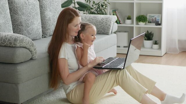 Beautiful mother and toddler daughter watch images on laptop. Mother and daughter spend time together at home. Cute small girl sits on knees of mom and and has fun with her.