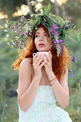 Tea party ceremony. Herbal medicine, health concept. A beautiful red-haired woman in a wreath of wildflowers is sitting on the grass and drinking natural herbal tea. Light breakfast and diet in nature