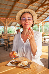 young woman in glasses and a hat eats sea fish cooked in a restaurant on the seaside in the summer