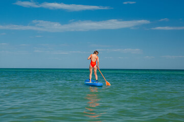 a young woman in a red swimsuit on the background of the sea stands on a surfing board holding a paddle in her hands