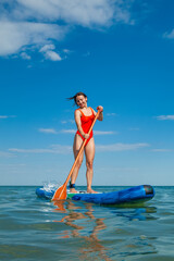 a young woman in a red swimsuit on the background of the sea stands on a surfing board holding a paddle in her hands