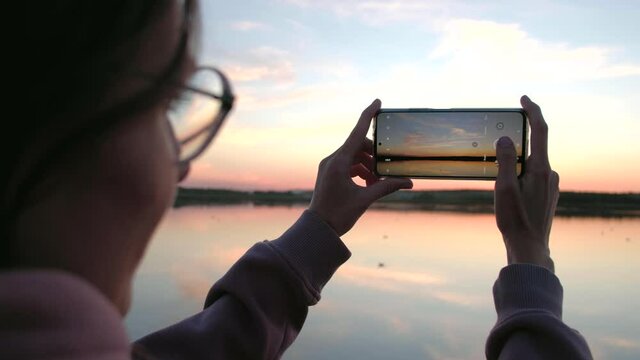 Young woman in glasses taking photo of sunset holding smartphone with your hands near the lake.