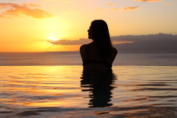 Fototapeta premium A girl in a swimsuit with her hair down stands back in the pool and looks at the sunset and the ocean.