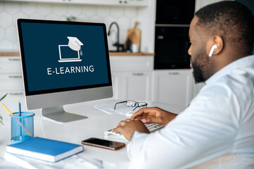 Online education, e-learning concept. African American smart confident guy sitting at home at work desk, uses computer, on computer screen the inscription e-learning. Study remotely
