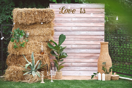 Rustic wedding photo zone decorated with wooden background, hay, wicker pots with plants, lamps and candles. Copy, empty space for text