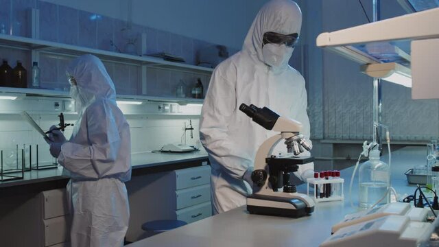 Slowmo shot of unrecognizable black male epidemiologist in disposable coveralls, goggles, gloves and face mask carrying rack with test tubes filled with blood samples, then using microscope in lab
